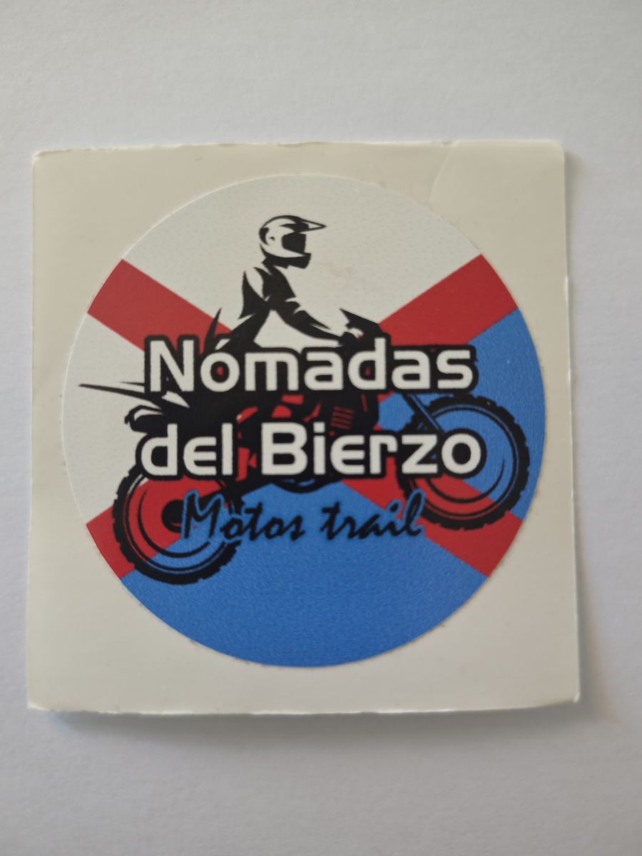 Logo original de Nómadas del Bierzo: adhesivo de vinilo antiguo y arrugado, fotografiado con el móvil sobre una mesa. El color está lavado y el papel doblado.