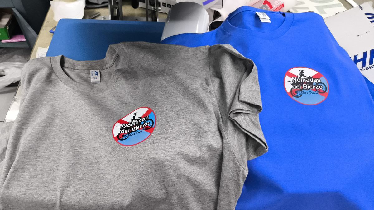 Camisetas gris y azul con el emblema circular de Nómadas del Bierzo impreso en el pecho mediante DTF.